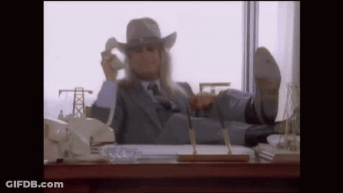 A Man In A Suit And Cowboy Hat Is Sitting At A Desk With His Feet Up Talking On A Cell Phone . GIF