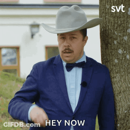 A Man In A Suit And Cowboy Hat Is Standing Next To A Tree And Says Hey Now GIF