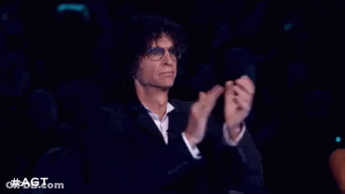 A Man In A Suit And Glasses Is Sitting In A Dark Room Clapping His Hands . GIF