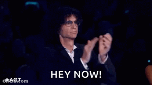 A Man In A Suit And Glasses Is Sitting In A Dark Room With His Hands In The Air . GIF