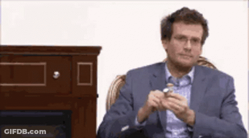 A Man In A Suit And Glasses Is Sitting In Front Of A Fireplace Holding Something In His Hand . GIF