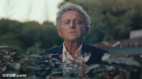 A Man In A Suit And Glasses Is Standing In Front Of A Bush With His Eyes Closed GIF