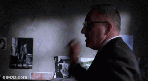 A Man In A Suit And Glasses Is Standing In Front Of A Wall With Photos On It GIF