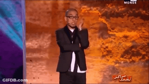 A Man In A Suit And Glasses Stands On A Stage With The Word Monde On The Bottom Left GIF
