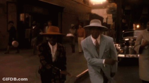 A Man In A Suit And Hat Is Dancing With A Woman In A Dress On A City Street . GIF