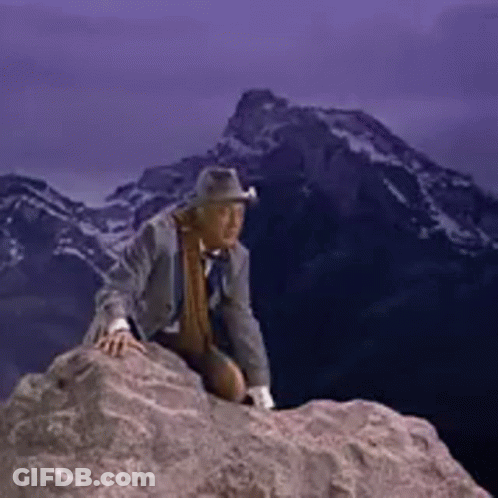 A Man In A Suit And Hat Is Sitting On Top Of A Rock . GIF