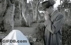A Man In A Suit And Hat Is Smoking A Cigarette While Standing Next To A Body . GIF