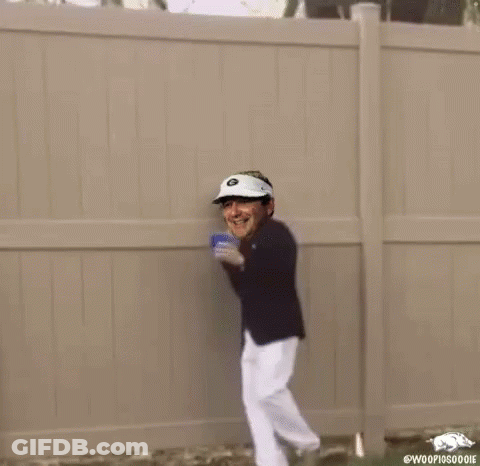 A Man In A Suit And Hat Is Standing In Front Of A Fence Holding A Cup . GIF