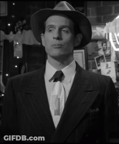 A Man In A Suit And Hat Is Standing In Front Of A Wall With Pictures GIF