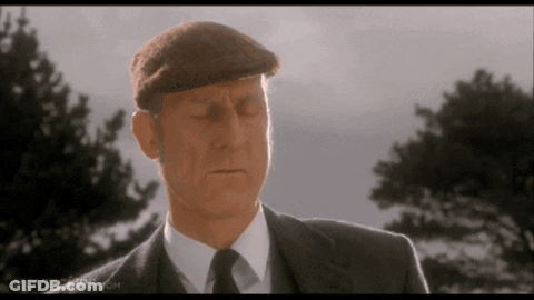 A Man In A Suit And Hat Is Standing In Front Of Some Trees . GIF