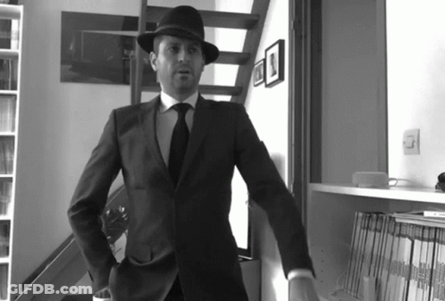 A Man In A Suit And Hat Stands In Front Of A Bookshelf With Magazines On It GIF
