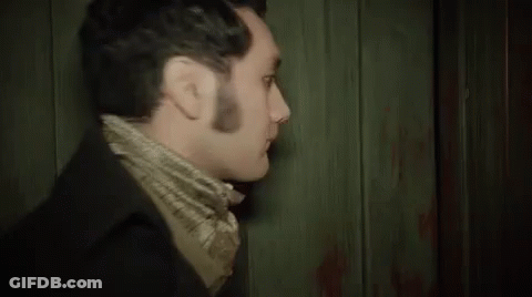 A Man In A Suit And Scarf Is Standing In Front Of A Wooden Wall And Looking At The Camera . GIF