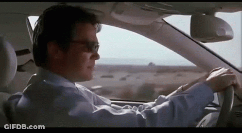 A Man In A Suit And Sunglasses Is Driving A Car On A Highway . GIF