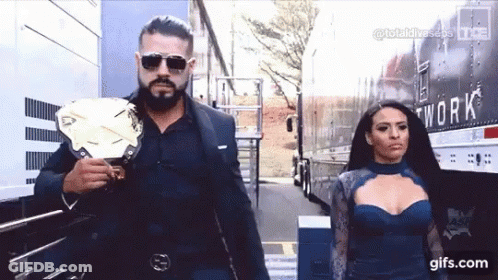 A Man In A Suit And Sunglasses Is Holding A Wrestling Championship Belt Next To A Woman In A Blue Dress . GIF