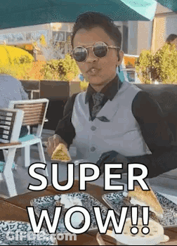 A Man In A Suit And Sunglasses Is Sitting At A Table Eating A Sandwich . GIF