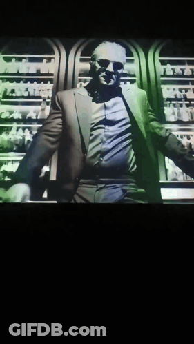 A Man In A Suit And Sunglasses Is Standing In Front Of A Display Of Bottles GIF