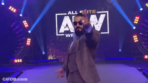 A Man In A Suit And Sunglasses Is Standing In Front Of A Sign That Says Wrestling Saturday Night Dynamite GIF