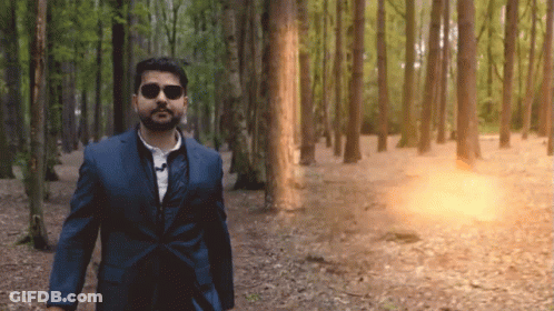 A Man In A Suit And Sunglasses Is Walking Through The Woods GIF