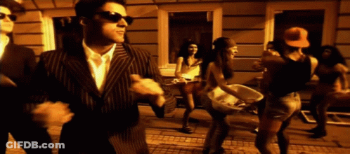 A Man In A Suit And Sunglasses Stands In Front Of A Group Of People Dancing GIF