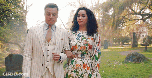 A Man In A Suit And Tie And A Woman In A Floral Dress Are Walking In A Park . GIF