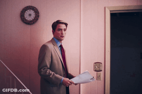 A Man In A Suit And Tie Holds A Piece Of Paper In Front Of A Pink Wall GIF
