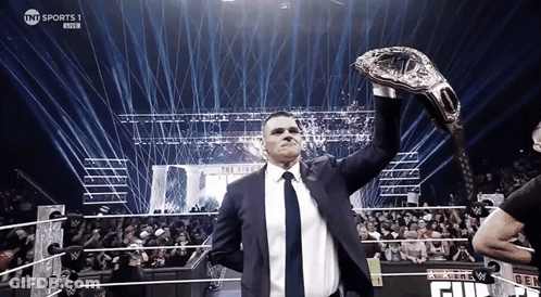 A Man In A Suit And Tie Holds Up A Wrestling Championship Belt In Front Of A Crowd GIF