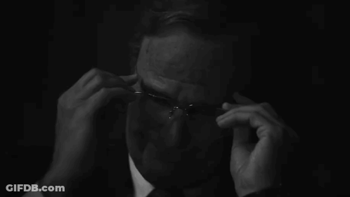 A Man In A Suit And Tie Is Adjusting His Glasses In The Dark . GIF
