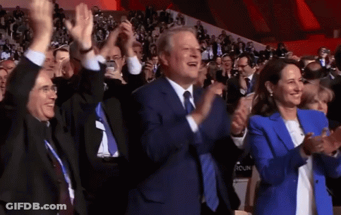 A Man In A Suit And Tie Is Applauding While A Woman In A Blue Jacket Looks On GIF