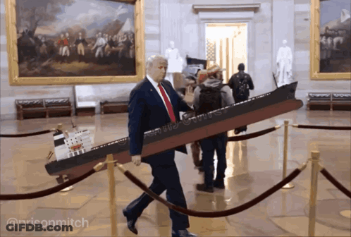 A Man In A Suit And Tie Is Carrying A Model Of A Ship That Says ' Donald Trump ' On It GIF
