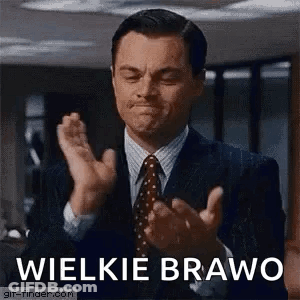 A Man In A Suit And Tie Is Clapping His Hands In Front Of A Sign That Says Wielkie Bravo . GIF