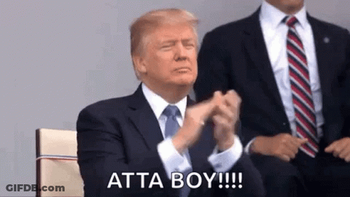 A Man In A Suit And Tie Is Clapping His Hands While Sitting In Front Of Another Man . GIF