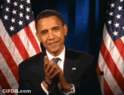 A Man In A Suit And Tie Is Clapping In Front Of American Flags . GIF