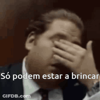 A Man In A Suit And Tie Is Covering His Face With His Hand And Says `` So Podem Estar A Brincar '' . GIF