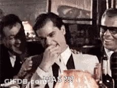 A Man In A Suit And Tie Is Covering His Mouth With His Hand While Sitting At A Table . GIF