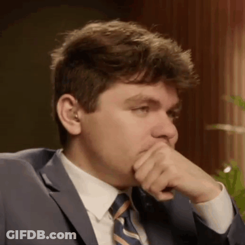 A Man In A Suit And Tie Is Covering His Mouth With His Hand While Sitting In A Chair . GIF