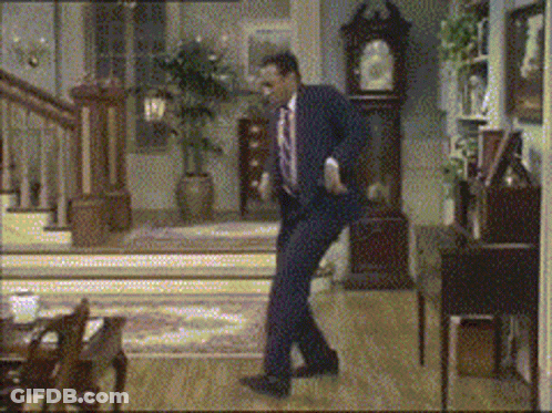 A Man In A Suit And Tie Is Dancing In A Living Room With A Clock In The Background GIF