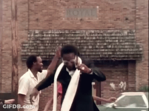 A Man In A Suit And Tie Is Dancing In Front Of A Brick Building With A Car In The Background . GIF