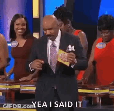 A Man In A Suit And Tie Is Dancing On A Game Show While A Group Of Women Are Standing Behind Him . GIF