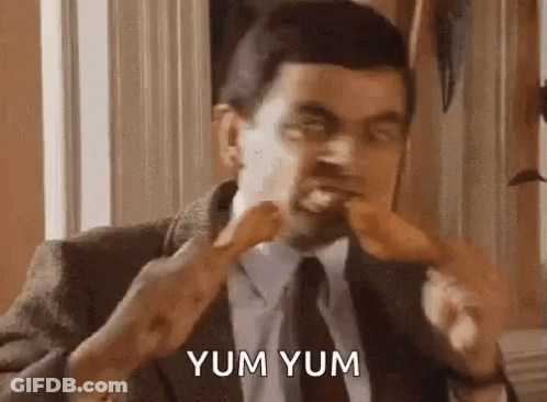 A Man In A Suit And Tie Is Eating A Chicken Leg And Saying Yum Yum . GIF