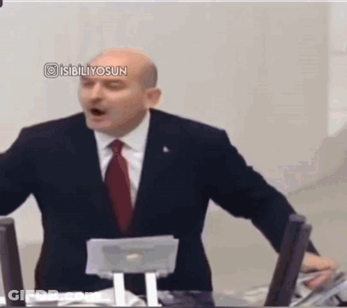 A Man In A Suit And Tie Is Giving A Speech In Front Of A Computer . GIF