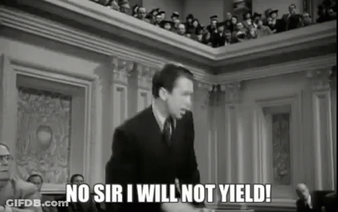 A Man In A Suit And Tie Is Giving A Speech In Front Of A Crowd Of People In A Courtroom . GIF