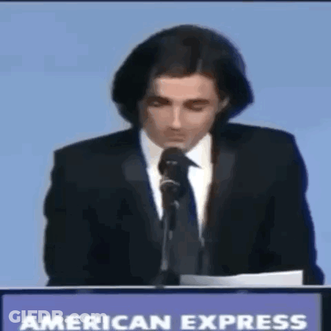 A Man In A Suit And Tie Is Giving A Speech In Front Of An American Express Sign GIF