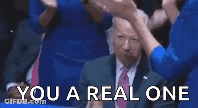 A Man In A Suit And Tie Is Giving A Thumbs Up While Sitting In A Crowd Of People . GIF