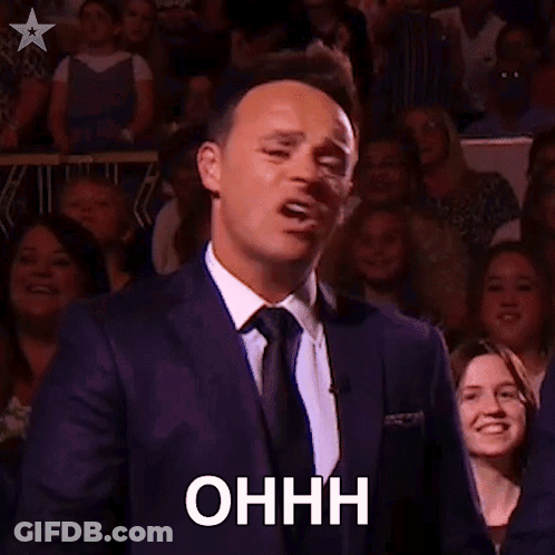 A Man In A Suit And Tie Is Holding A Microphone In Front Of A Crowd And Making A Funny Face . GIF