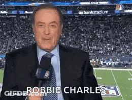 A Man In A Suit And Tie Is Holding A Microphone While Standing In Front Of A Football Field . GIF