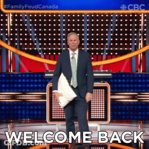 A Man In A Suit And Tie Is Holding A Pillow In Front Of A Stage That Says Welcome Back GIF