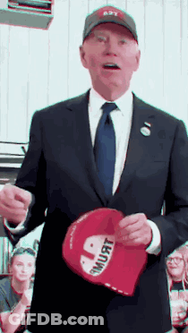 A Man In A Suit And Tie Is Holding A Red Hat That Says " I Must " GIF