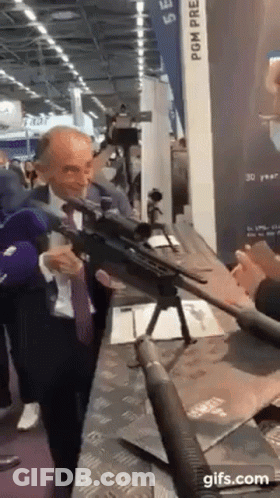 A Man In A Suit And Tie Is Holding A Rifle In Front Of A Camera . GIF