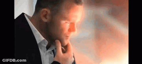 A Man In A Suit And Tie Is Holding His Hand To His Chin And Looking Down . GIF