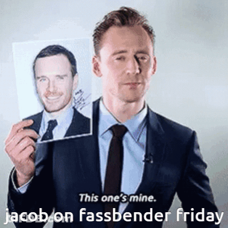 A Man In A Suit And Tie Is Holding Up A Picture Of Jacob On Fassbender Friday GIF